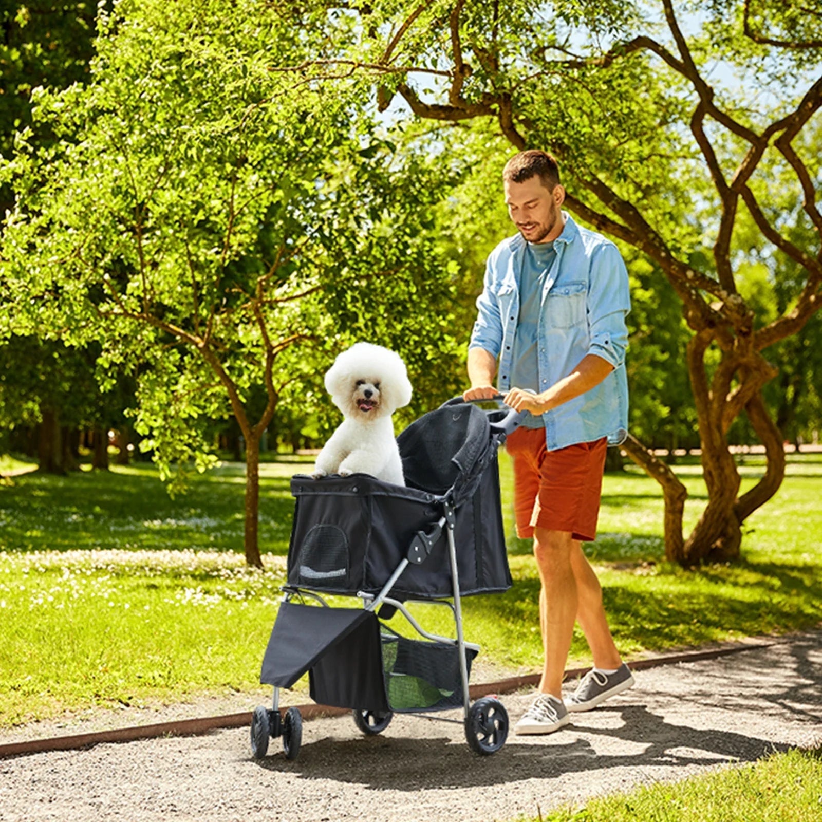 All Dogs Love 3-wheel foldable pet stroller with a white dog in a park, pushed by a man on a sunny day.