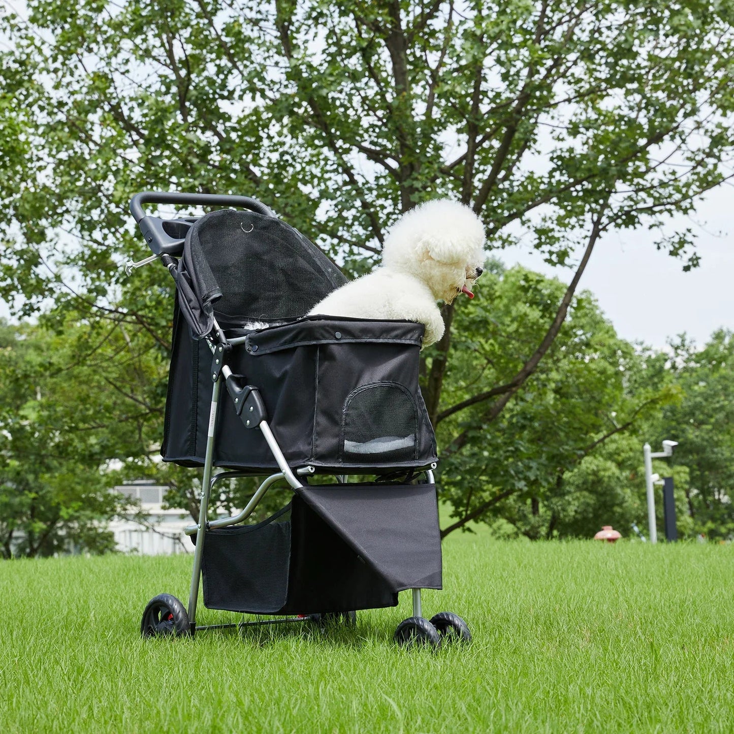 All Dogs Love foldable 3-wheel pet stroller in black with a white Bichon Frise sitting in the mesh cabin in a park